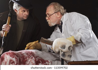 Grave Robber And Evil Doctor With Bloody Cleaver Exchange Conspiratorial Glances. Stage Effect, Isolated On Black Background, Spot Lighting.