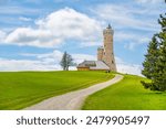 A gravel path leads to the Dalimil Lookout Tower in the Czech Republic. The tower stands tall against a backdrop of lush green grass and a blue sky with fluffy clouds.
