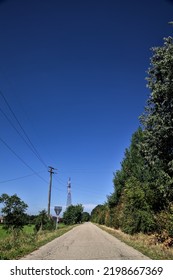 Gravel  Road Bordered By Fields With Electricity Poles At Its Edge And A Country House In The Distance On A Summer Day In The Italian Countryside