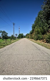 Gravel  Road Bordered By Fields With Electricity Poles At Its Edge And A Country House In The Distance On A Summer Day In The Italian Countryside