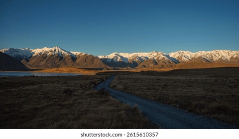 Gravel road leading to Southern Alps idyllic remote calm serene golden hour snow capped mountain peaks, Lake Heron winter sunset landscape, Ashburton Lakes Canterbury South Island New Zealand - Powered by Shutterstock - Get 15% off with code: PIKWIZARD15