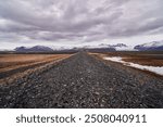 Gravel road is leading towards a mountain range with a dramatic sky in the background in iceland