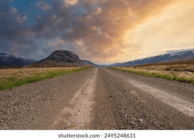 A gravel road stretches into the distance, flanked by grassy fields and leading towards a prominent mountain under a dramatic sky. Snow-capped mountains are visible in the background - Powered by Shutterstock