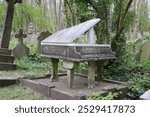 A gravestone designed as a grand piano with a raised lid, set amidst lush greenery and traditional headstones in Highgate Cemetery, London.