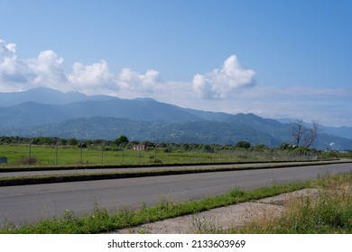 gray asphalt road behind which you can see the airport fence where grass grows along the edges and in the distance there are a number of mountain scenery