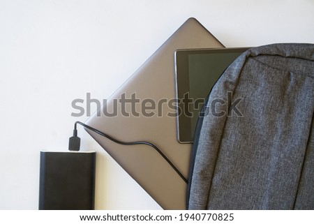 Gray backpack with black tablet and silver laptop or ultrabook lies on white background next to large capacious power bank. The concept of recharging gadgets while traveling or trip. View from above