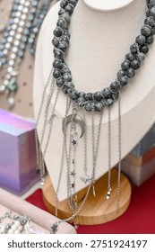 Gray beaded statement necklace with silver star charms and chains displayed on a jewelry bust, surrounded by colorful boxes and other accessories