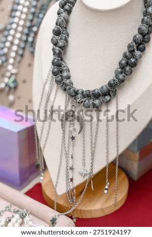 Gray beaded statement necklace with silver star charms and chains displayed on a jewelry bust, surrounded by colorful boxes and other accessories