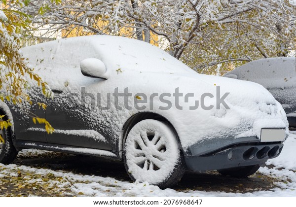 A gray car covered with the\
first snow against the background of yellow trees. First\
snowfall.