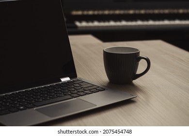 A gray cup of coffee next to a laptop on a wooden table against the background of a piano. Home Office. Horizontal view. Selective focus.