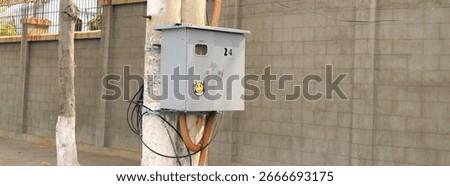 Gray electrical meter box with the number 24 and a smiley face sticker mounted on a concrete utility pole next to a block wall. Urban infrastructure and power supply concept.