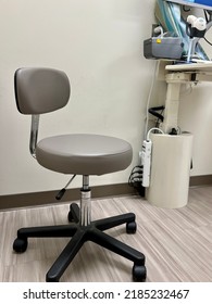 Gray, Generic And Nondescript Interior Of Medical Exam Room. Minimal Furniture Includes A Rolling Stool And Garbage Can.