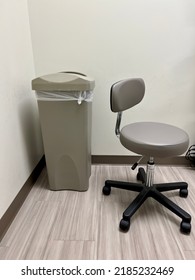 Gray, Generic And Nondescript Interior Of Medical Exam Room. Minimal Furniture Includes A Rolling Stool And Garbage Can.