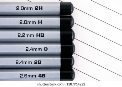 Gray Graphite Pencils With Graphite Grading Scale In A Row On Lined Paper Background.