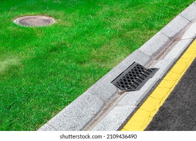 Gray Gutter Of A Stormwater Drainage System On The Side Of Asphalt Road With Yellow Markings And Sewer Septic Tank In Green Lawn, Concrete Drainage Ditch With Iron Grate.