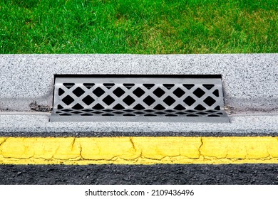 Gray Gutter Of A Stormwater Drainage System On The Side Of Asphalt Road With Yellow Markings And Green Lawn In Dry Weather, Concrete Drainage Ditch With Iron Grate Close-up Side View.