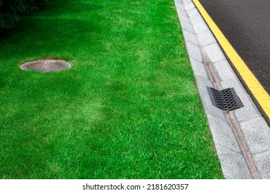 Gray Gutter Of A Stormwater System On The Side Of Asphalt Road With Yellow Markings And Manhole In Green Trimmed Lawn, Concrete Drainage Ditch With Iron Grate On Sunny Dry Weather, Nobody.