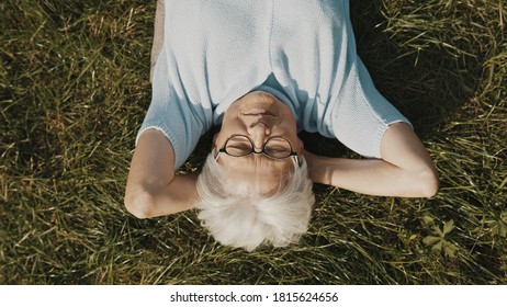 Gray Haired Woman Pensioner Lying On The Grass With Hands Under Her Head. Overhead Close Up Shot. High Quality Photo