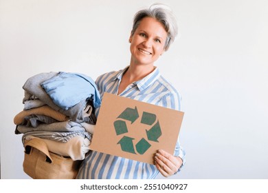 Gray haired woman with pile of clothes and recycling reusing signboard isolated on white background. Portrait mid age senior female posing with clothing stack made by eco friendly zero waste material - Powered by Shutterstock