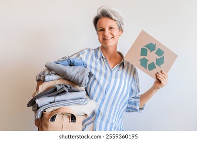 Gray haired woman with pile of clothes and recycling reusing signboard isolated on white background. Portrait mid age senior female posing with clothing stack made by eco friendly zero waste material - Powered by Shutterstock