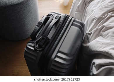 Gray hard shell suitcase stands. Black handle rests on top. Closed zipper secures travel belongings. White bedding touches luggage edge. Bedroom packing background suits vacation preparation.