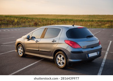 Gray hatchback car parked in an empty parking lot during sunset. Rear side exterior view of the vehicle outdoors with a scenic field background.