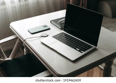 Portátil gris 2020, smartphone azul con carga inalámbrica sobre una mesa blanca de madera en la oficina con un portátil y gafas. Lugar de trabajo minimalista. Trabajar en casa. Coronavirus de cuarentena. Tecnología