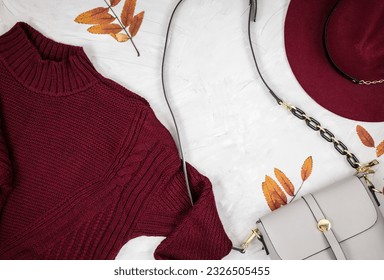 Gray leather woman bag, dark red sweater,  autumn leaves on gray background. Trendy autumn accessories. Cozy outfit