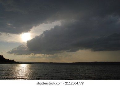 Gray menacing clouds over the river. The clouds are gray-white with a blue tint in the sky. The sun's rays shine through them. Below is a wide river and a bank overgrown with forest.
