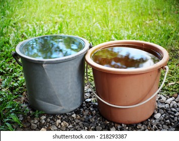 gray and orange plastic bucket on the floor outdoor surrounding with green area environment with reflection on water surface in the bucket showing leaves and tree silhouette and blue sky reflections - Powered by Shutterstock