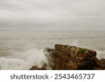 A gray, overcast sky above a rocky shore with ocean waves crashing.