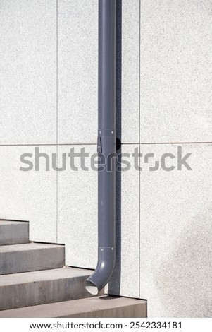 A gray pipe is attached to a building. The pipe is attached to a wall and is leaning against a wall