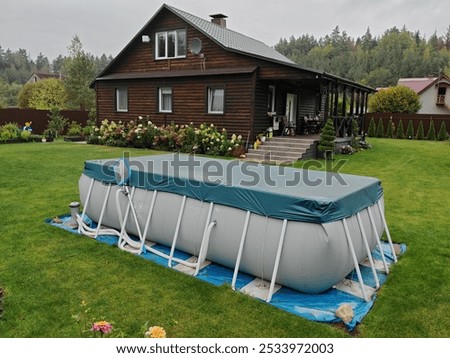 The gray PVC frame rectangular pool is located on a grassy lawn and is covered with an awning. A wooden cottage stands nearby, thuja trees and flowers grow. Overcast autumn weather and gray sky
