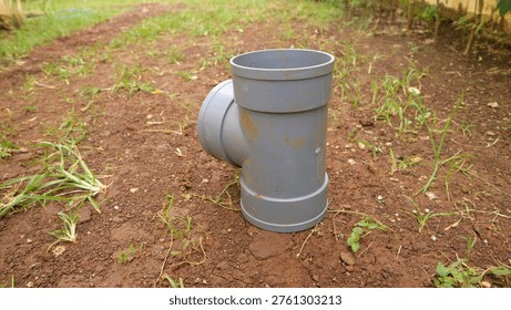 A Gray pvc pipe fitting elbow joint standing on soil outdoors in garden yard, plumbing drainage connector on ground with grass and dirt background.