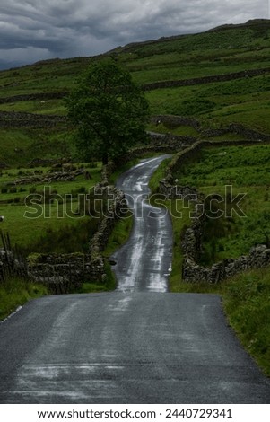 Gray rain clouds over a wet asphalt road winding through green hilly landscape. Moody weather while exploring and travelling around beautiful and peaceful mountainous countryside in Lake District.