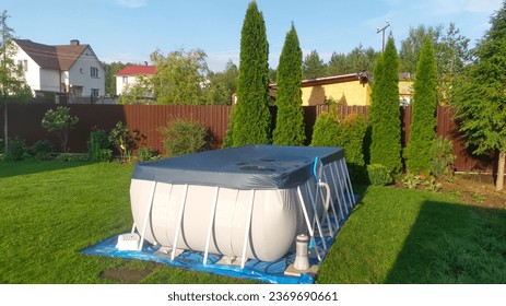 A gray rectangular frame pool stands on a grassy lawn and is covered by a shade. Near the metal fence there are thuja trees and flowers in flowerbeds. The pool is made of PVC on a plastic frame