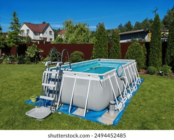 The gray rectangular frame pool stands on a grassy lawn. The water is filtered and there are toys floating in it. There are thuas and flowers growing next to the metal fence. The pool is made of PVC