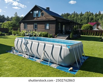 A gray rectangular frame pool stands on the grassy lawn next to the cottage. The water is filtered. Thuja trees and flowers grow next to the metal fence. The pool is made of PVC. Sunny summer weather