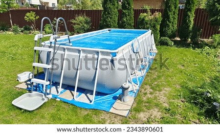 The gray rectangular frame pool stands on a grassy lawn. The surface of the water is covered with a floating awning. Nearby there are thuas and flowers in flowerbeds. The pool is made of PVC