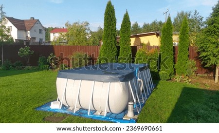 A gray rectangular frame pool stands on a grassy lawn and is covered by a shade. Near the metal fence there are thuja trees and flowers in flowerbeds. The pool is made of PVC on a plastic frame