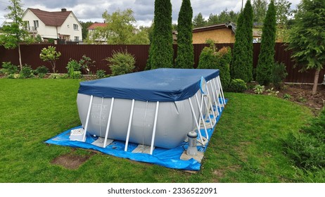 A gray rectangular frame swimming pool stands on the grassy lawn of the infield and is covered with an awning. Nearby thuja and flowers grow in flower beds The pool is made of PVC material