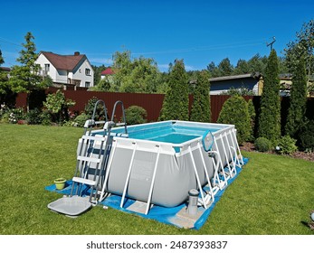 The gray rectangular frame swimming pool stands on a grassy lawn. The water is cleaned with a filter. Next to the metal fence there are thuja trees and flowers. The pool is made of PVC. Sunny weather