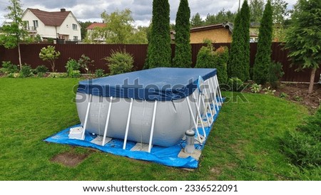 A gray rectangular frame swimming pool stands on the grassy lawn of the infield and is covered with an awning. Nearby thuja and flowers grow in flower beds The pool is made of PVC material