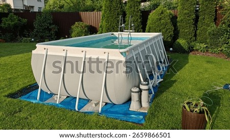 A gray rectangular PVC frame pool stands on a grassy lawn. Nearby is a filter and heater to clean and heat the water. Thuja trees, ornamental bushes and flowers grow next to the metal fence. Sunny