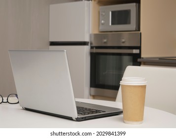 Gray Silver Laptop, Mobile Phone ,  Paper Coffee Cup Standing On White Wooden Table With Kitchen Background. No People.