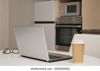 Gray Silver Laptop, Mobile Phone ,  Paper Coffee Cup Standing On White Wooden Table With Kitchen Background. No People.