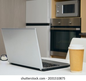 Gray Silver Laptop, Mobile Phone ,  Paper Coffee Cup Standing On White Wooden Table With Kitchen Background. No People.