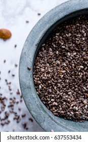 A Gray Stone Bowl Of Chia Seeds Overflowing Onto A White Marble Surface.