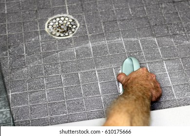 Gray tile shower floor being cleaned by an adult hand of a man with a scrub brush. Shower tile and grout being cleaned by the hand of a man with a scrub brush and soap