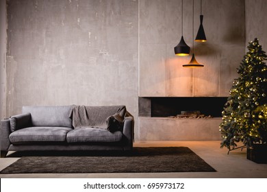 The gray velor sofa in the dark loft room has a bright light from the eternal light and an artificial fireplace. Inner attic with concrete walls and a decorated Christmas tree with gift boxes